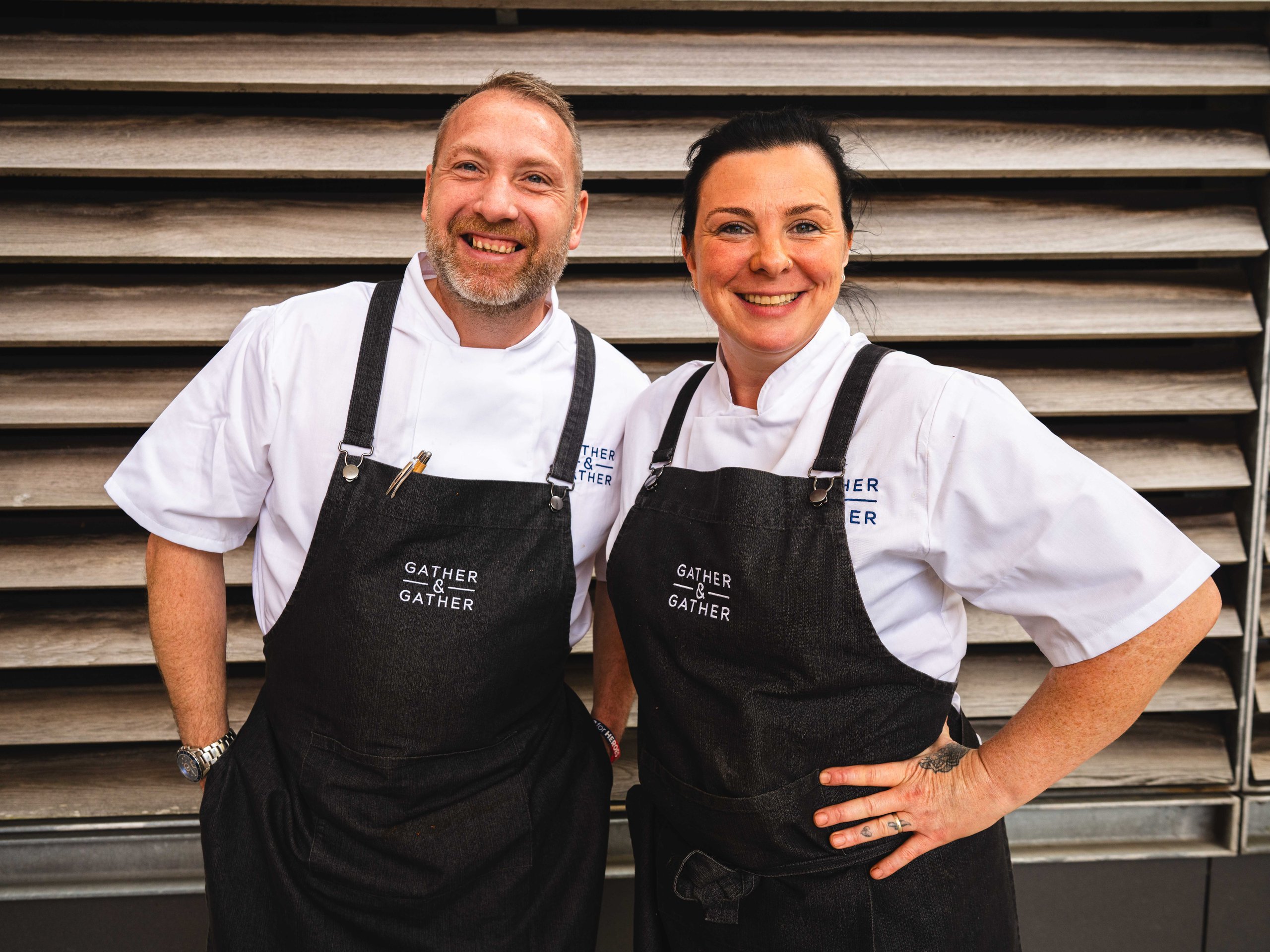 Two chefs wearing aprons, smiling, standing in front of a wooden background.