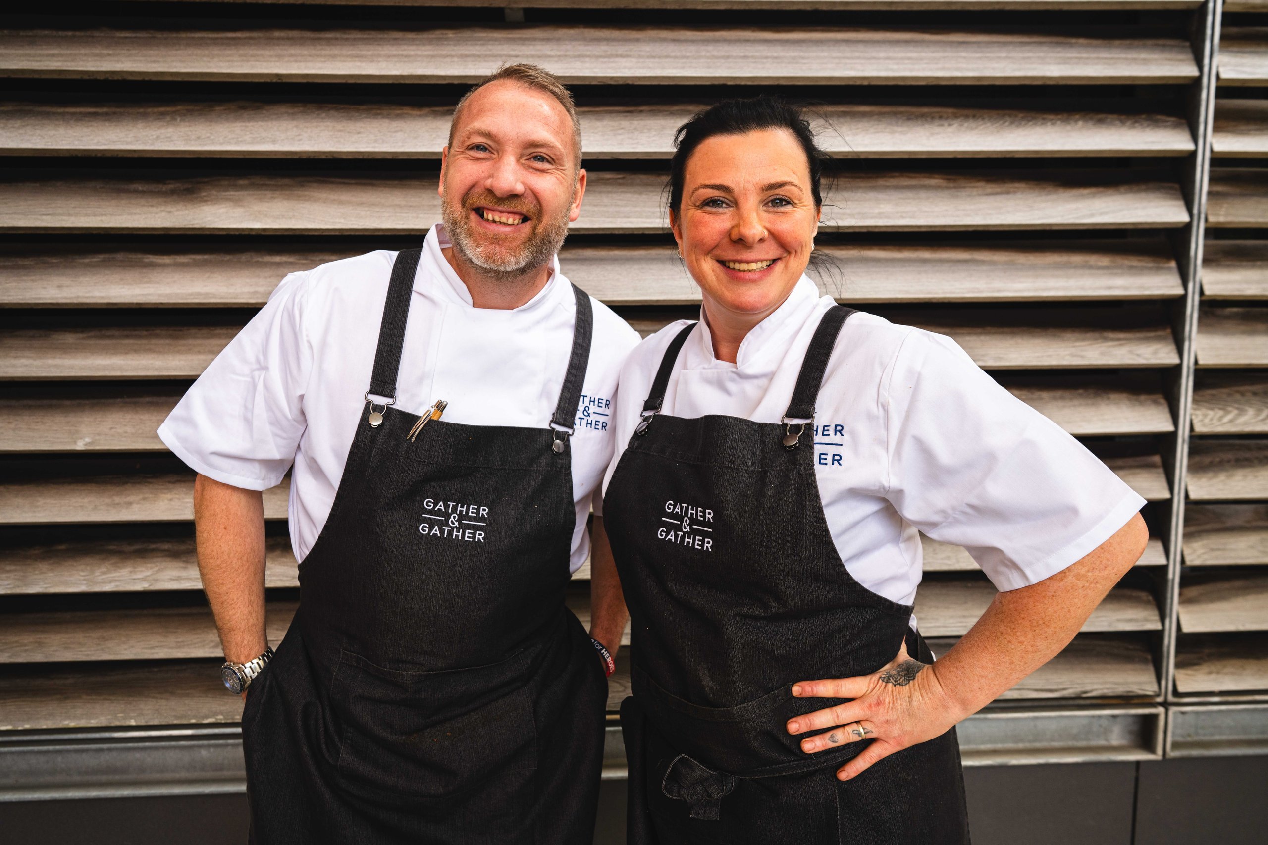 Two chefs wearing aprons, smiling, standing in front of a wooden background.