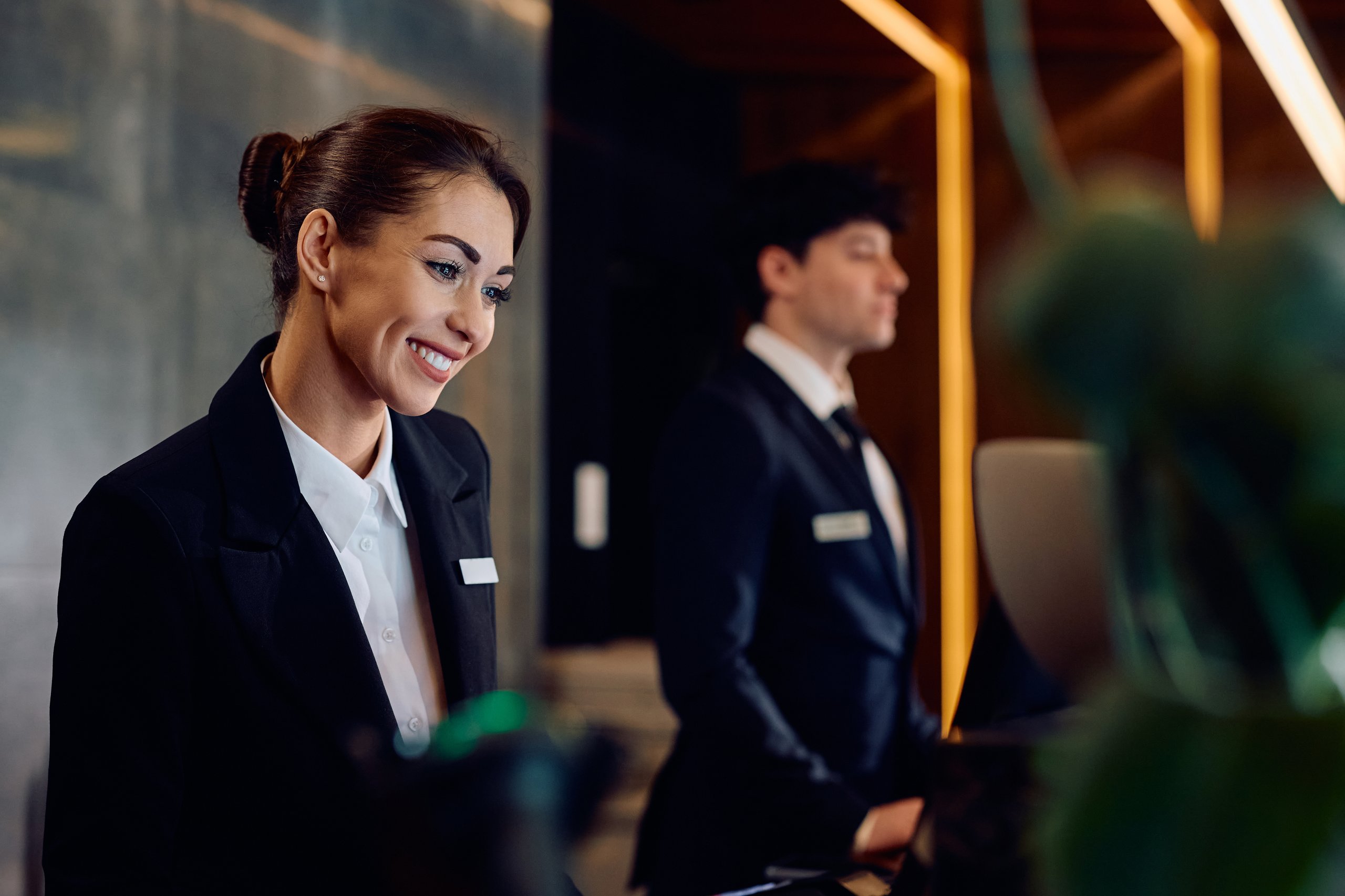 Hotel receptionist smiling, looking down. Employee wearing a name tag and blazer.