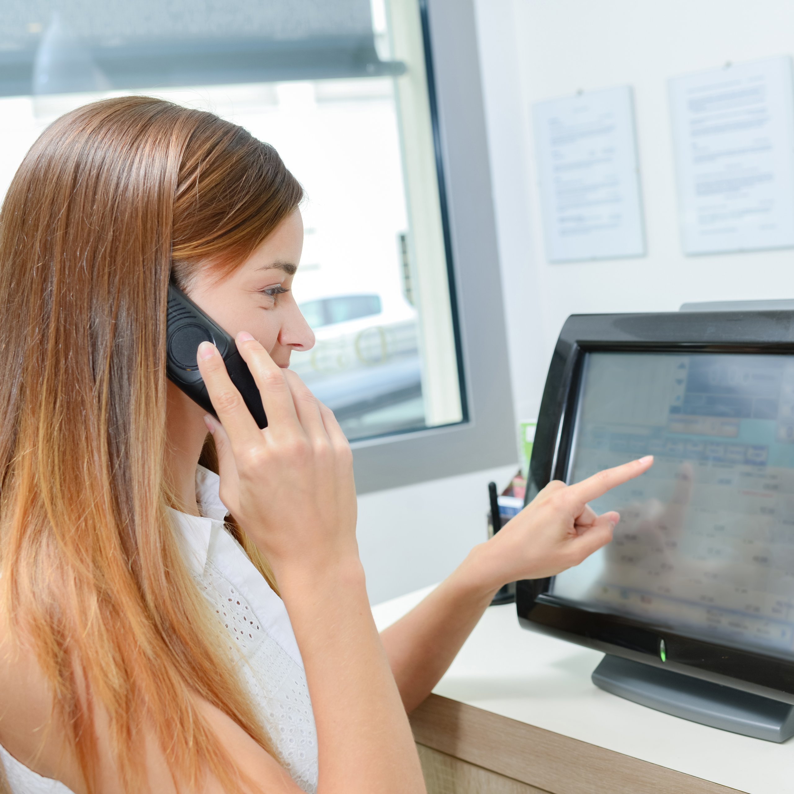Woman talking on the phone at a cash register, pointing at screen.