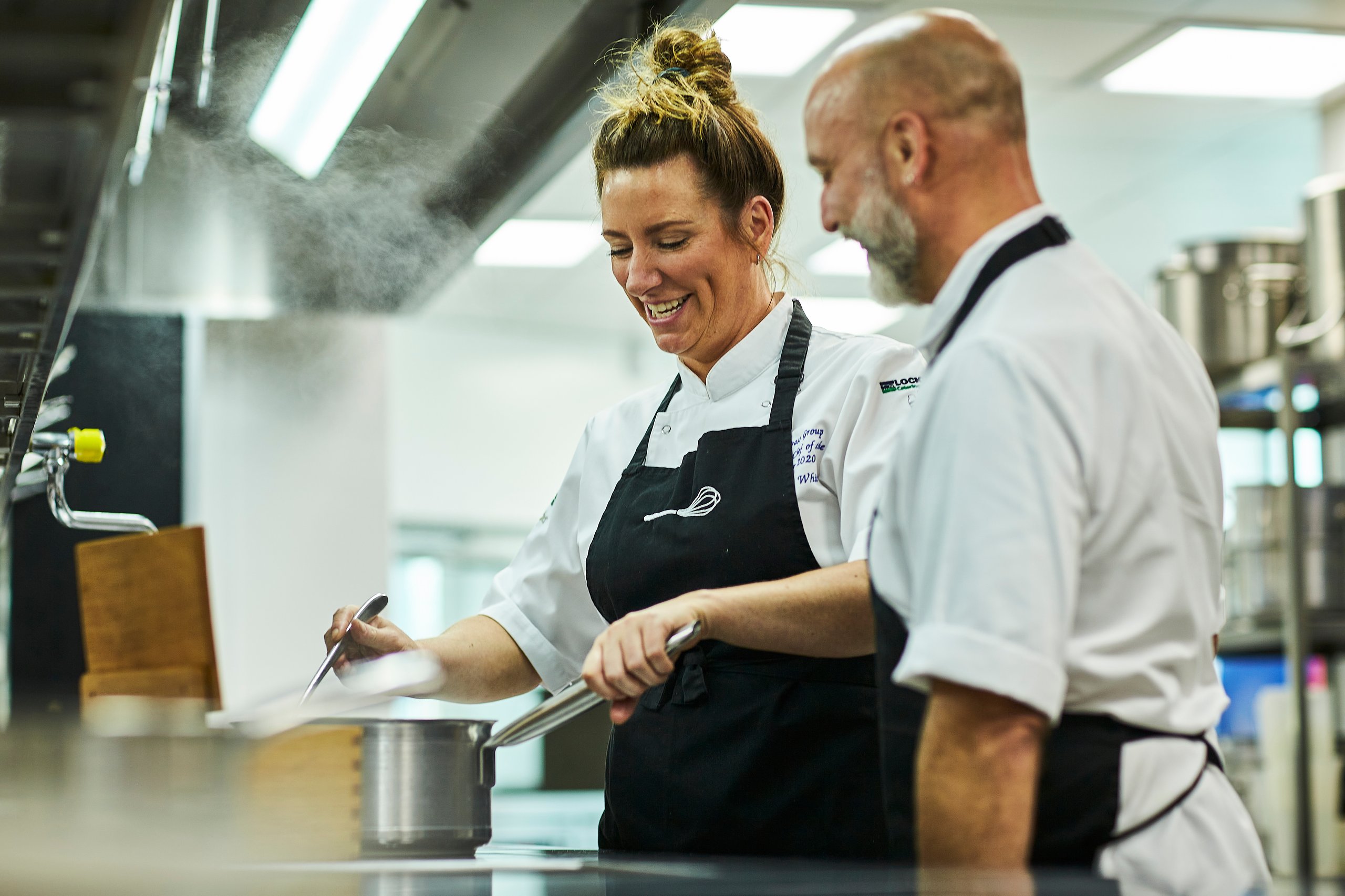 Chefs smiling, cooking in a professional kitchen, steam rising.