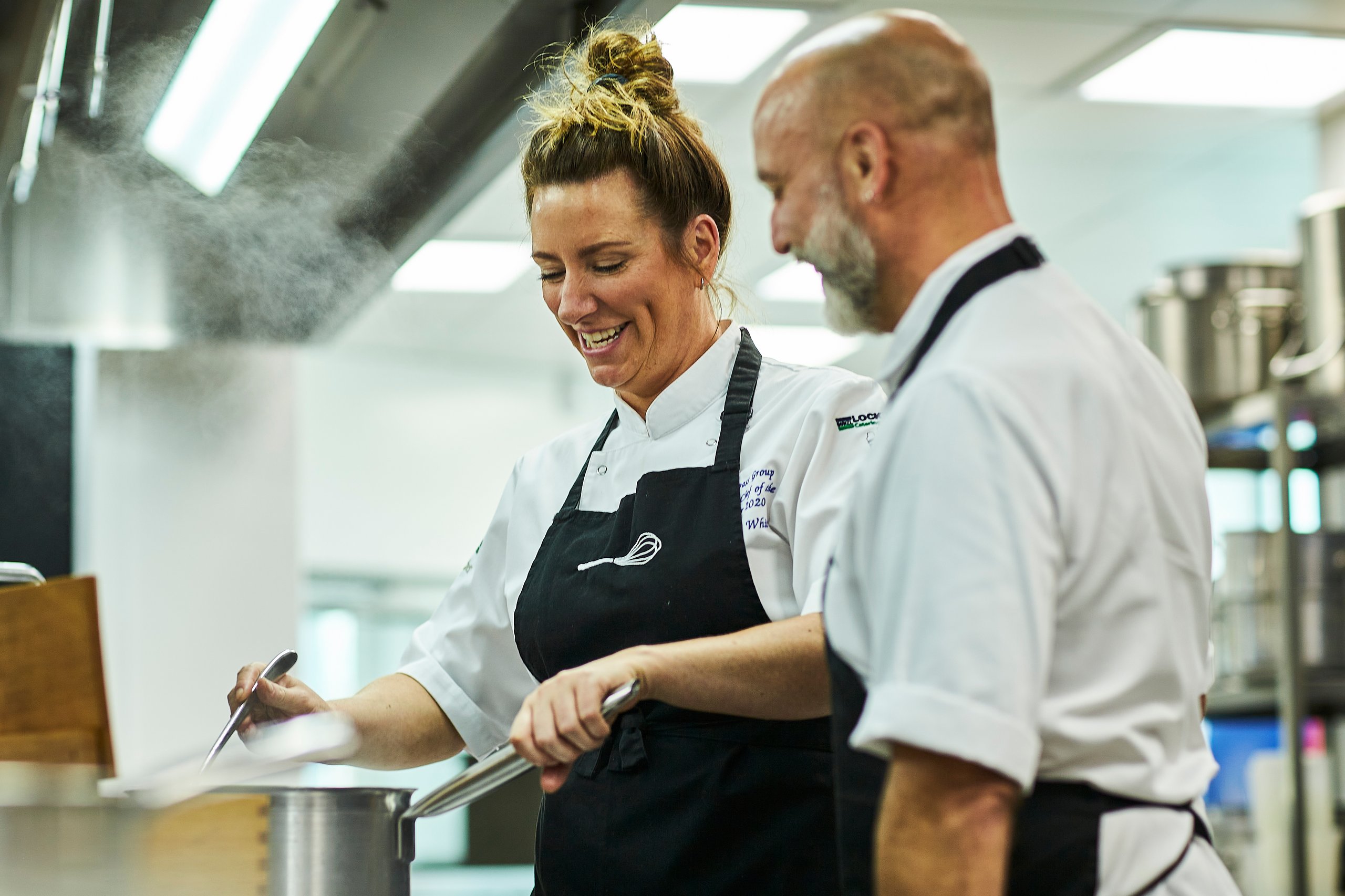 Chefs smiling, cooking in a professional kitchen, steam rising.