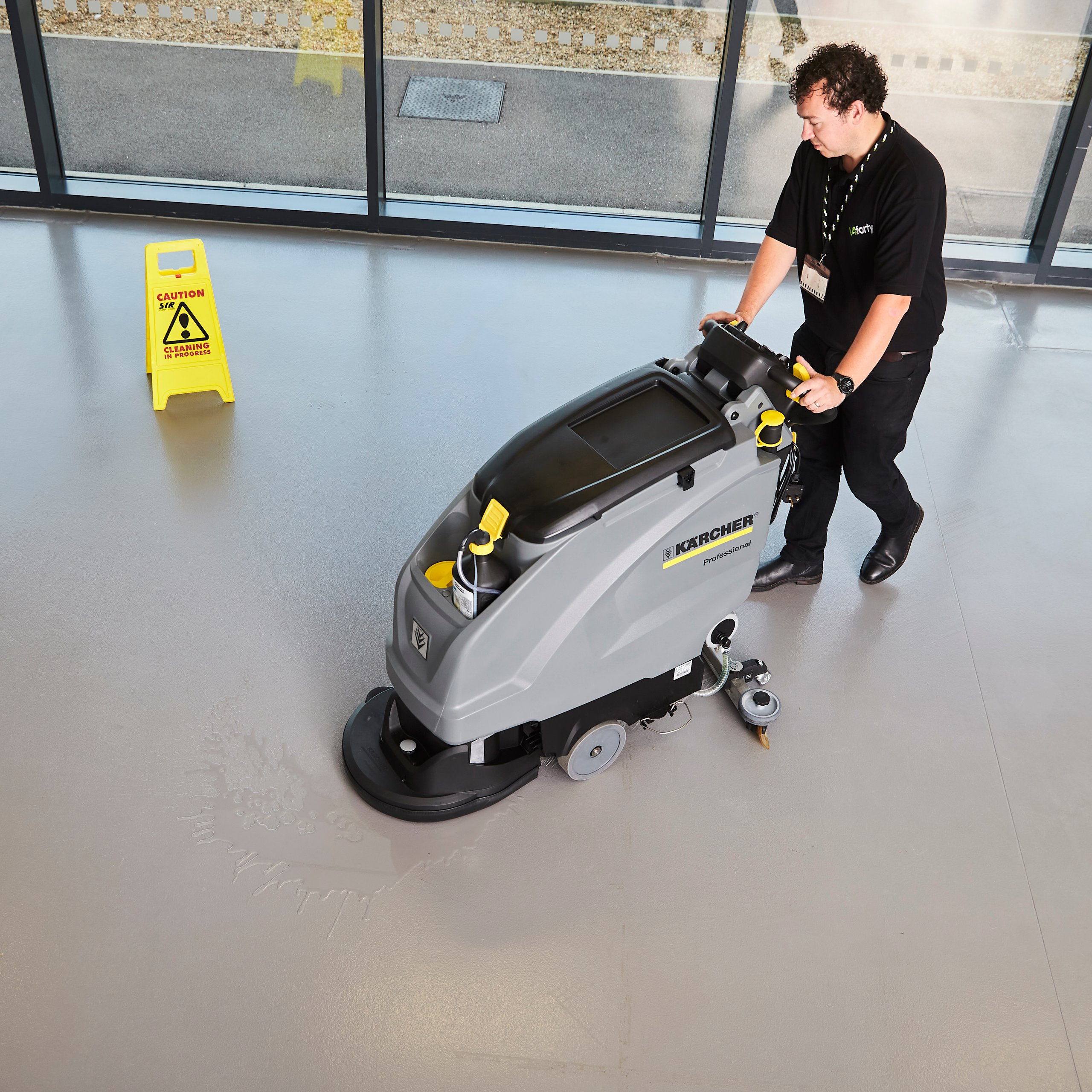 A man using a floor cleaner in an office. A "cleaning in progress" sign is near.