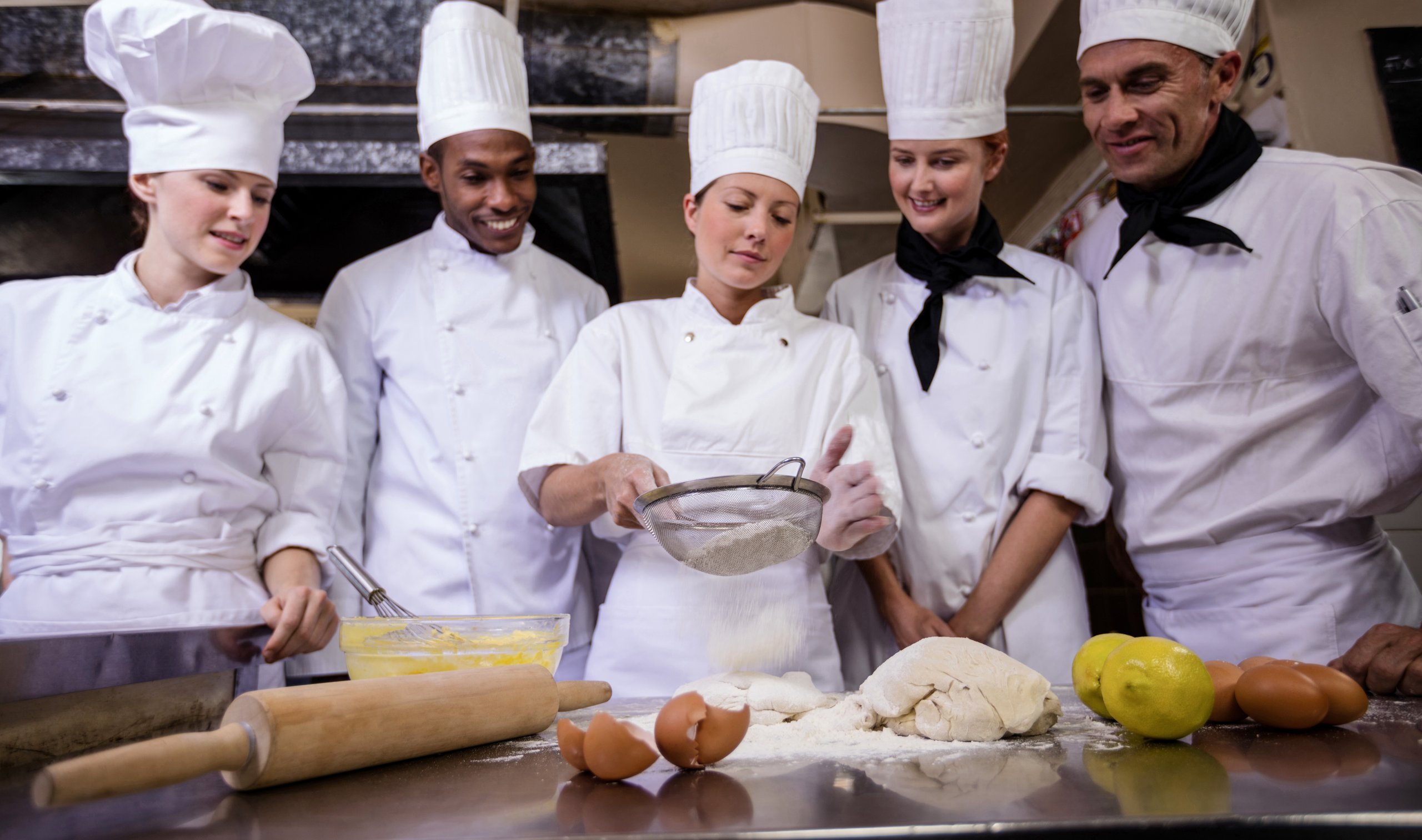 Five chefs in a kitchen are gathered around a table where one chef is sifting flour. Ingredients like lemons, eggs, and dough are visible.