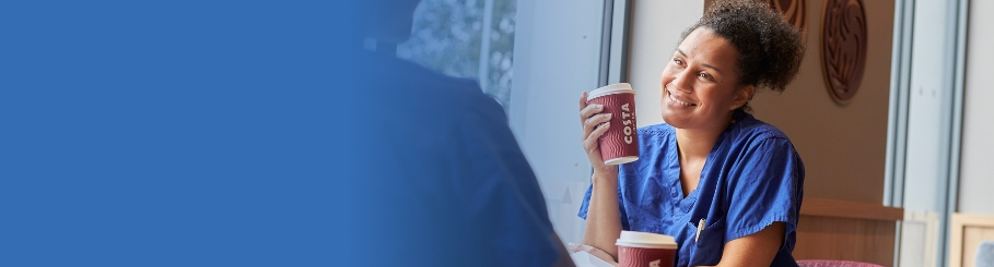 A nurse in scrubs smiles while holding a Costa Coffee cup. Another cup rests nearby.