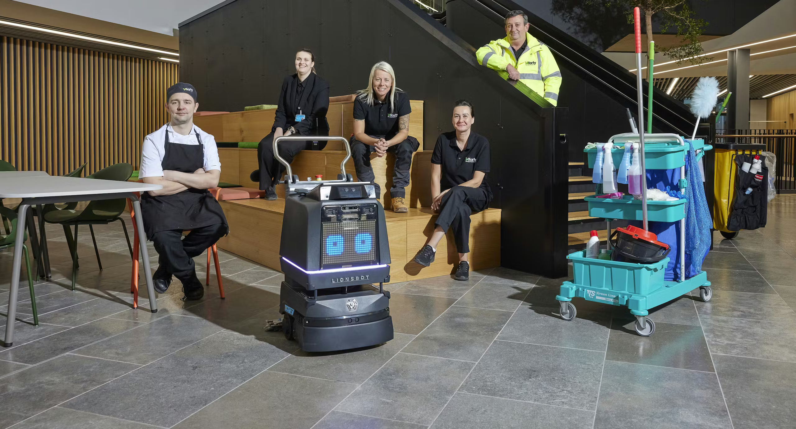 A team of five people and a cleaning robot are in a modern office building. A cleaning cart is nearby.