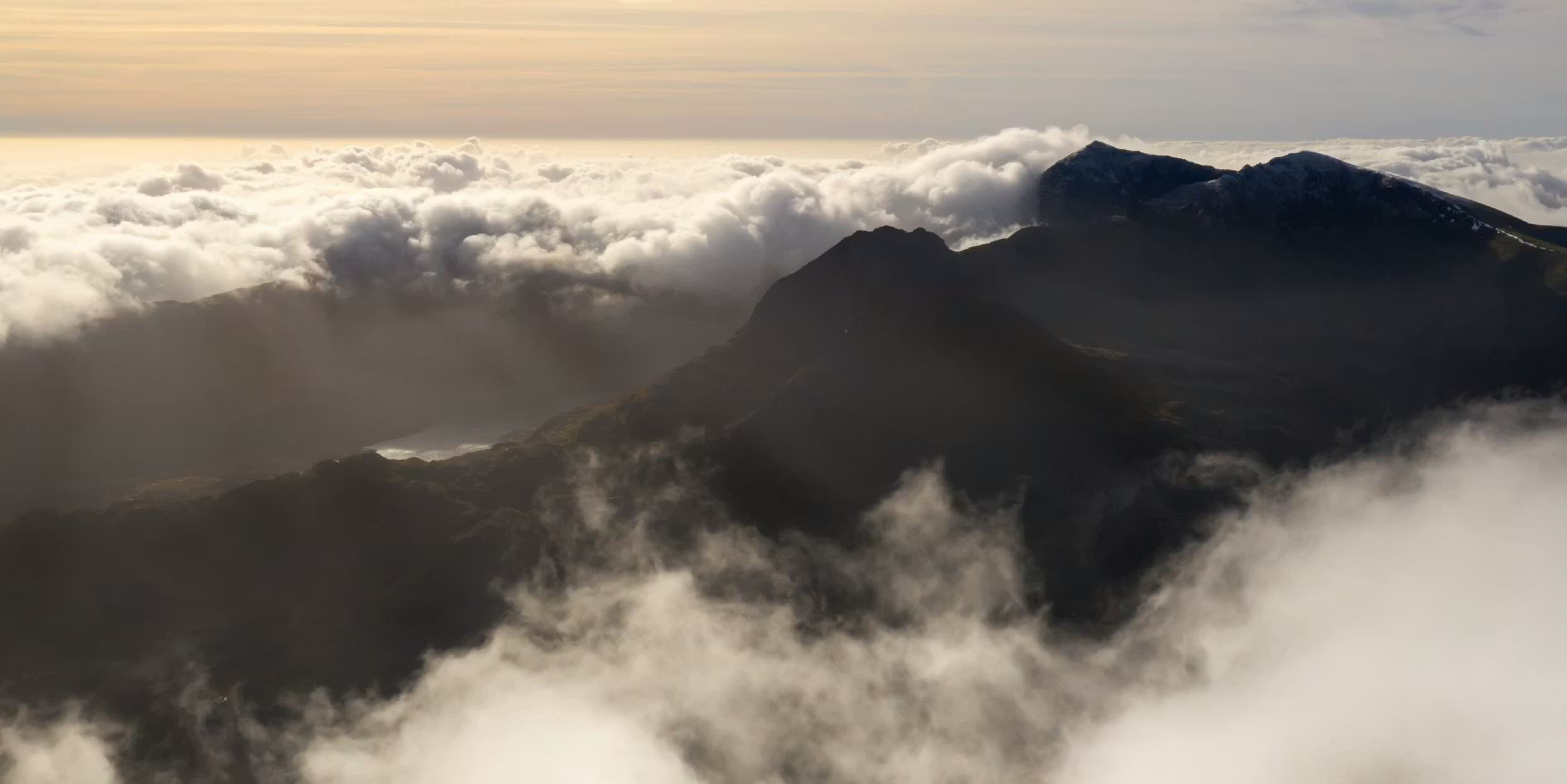 Silhouettes of dark mountain peaks emerge from dramatic cloud formations under a soft, golden sky.