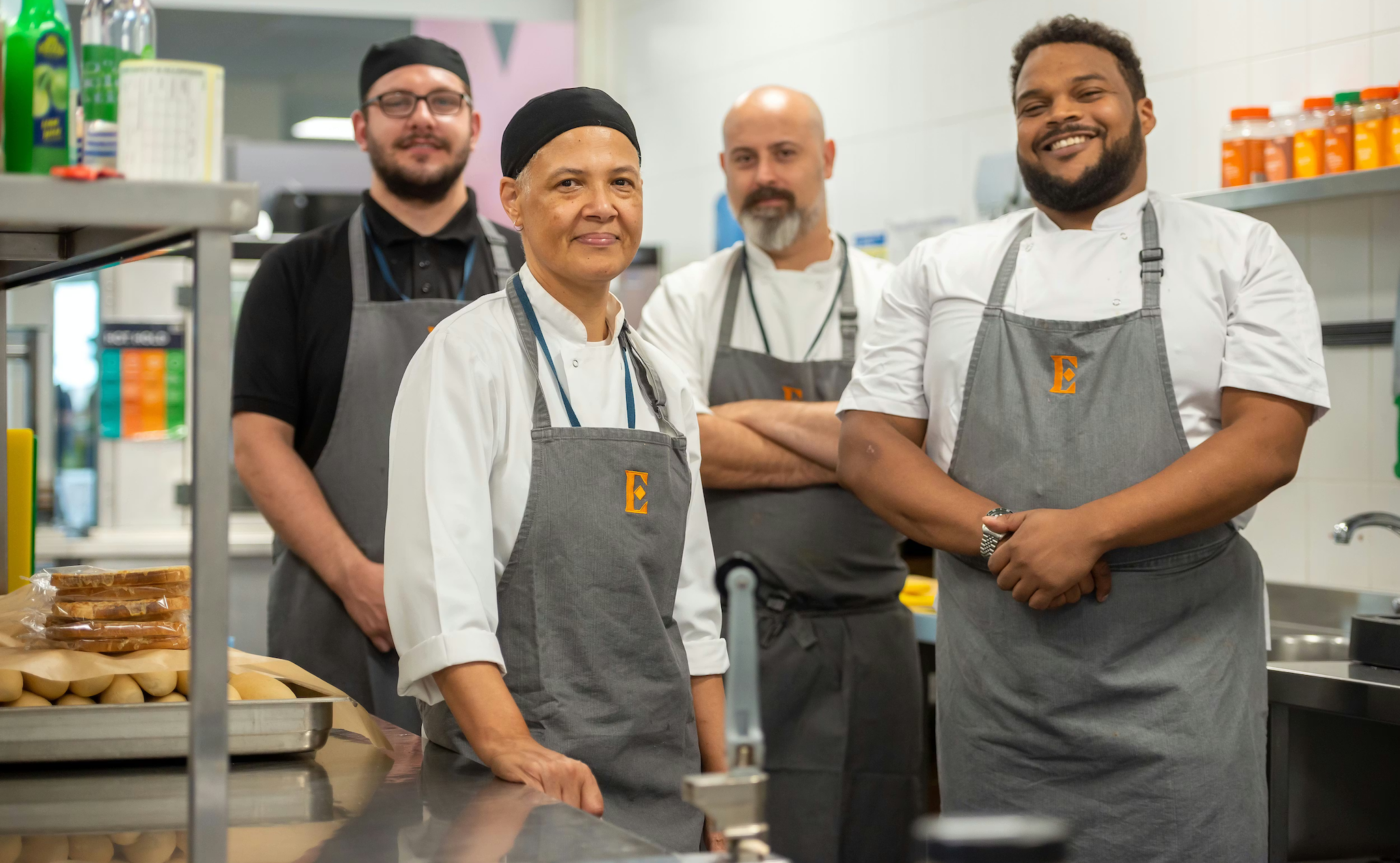 Four chefs in grey aprons with an orange "E" logo stand in a commercial kitchen. They are smiling at the camera.