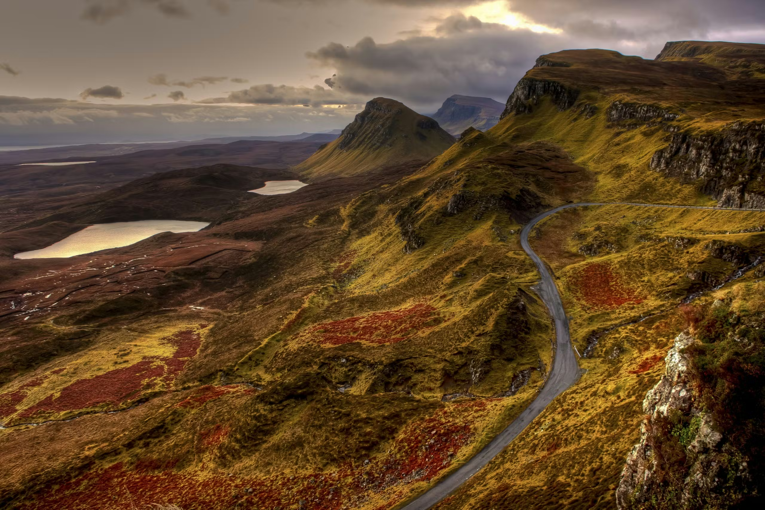 A winding road cuts through a rugged Scottish Highlands landscape with rugged hills, two lakes, and dramatic, cloudy skies overhead.
