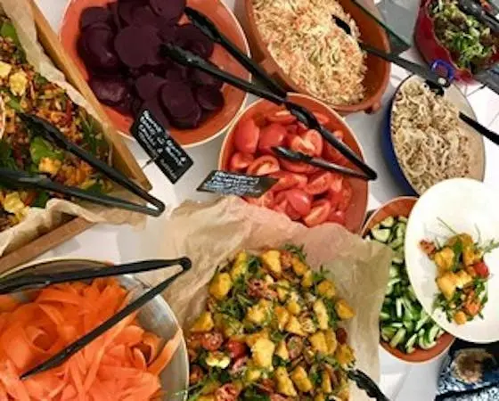 A variety of colorful salads and chopped vegetables are displayed in bowls on a table, with tongs resting in each bowl.