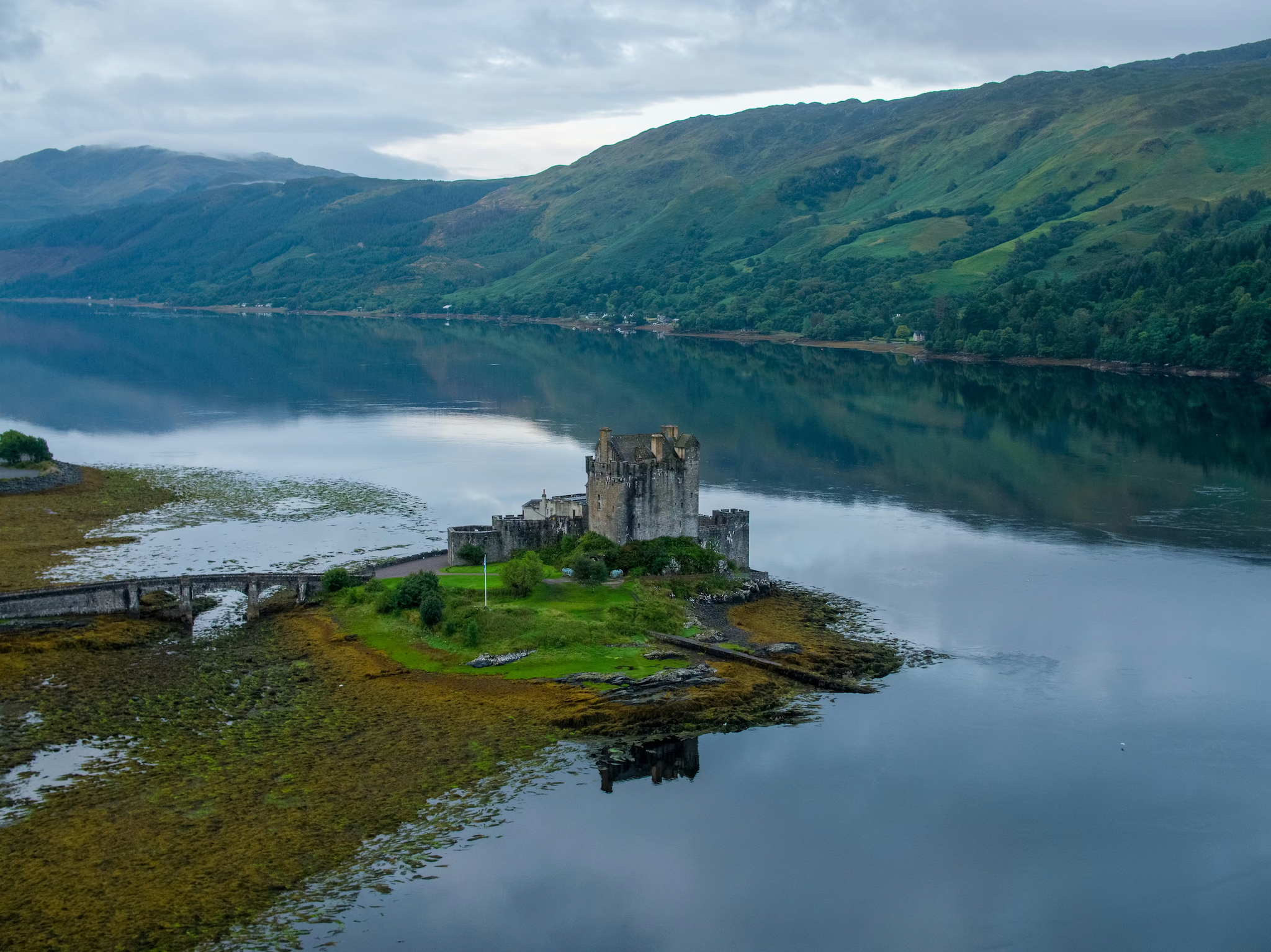 Eilean Donan Castle sits on a small island, connected by a stone bridge, surrounded by calm water and hills.