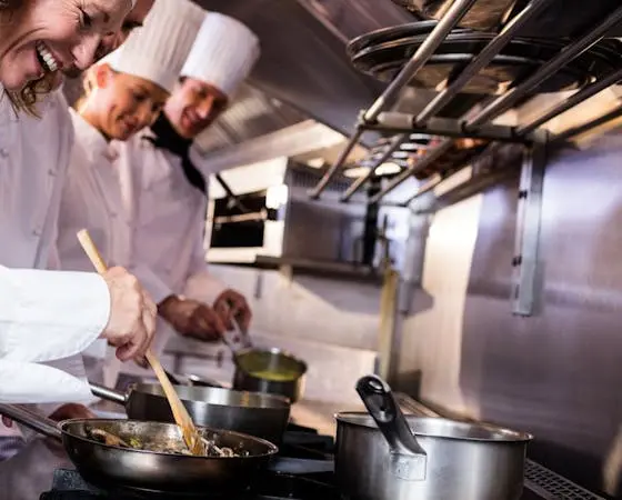 Three chefs in a busy commercial kitchen preparing food, with one stirring ingredients in a pan and others looking on.