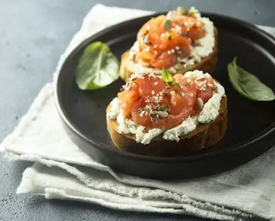 Two crostini topped with cream cheese, smoked salmon, seeds, and fresh herbs on a dark plate with a napkin.