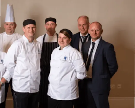 A group of chefs and two men in suits pose for a photo. The chefs wear white uniforms and black caps.
