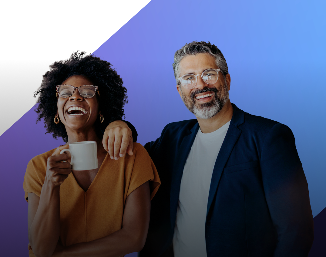 Two colleagues, a Black woman with glasses holding a mug and laughing, and a white man with glasses and a beard, stand together with his arm around her.
