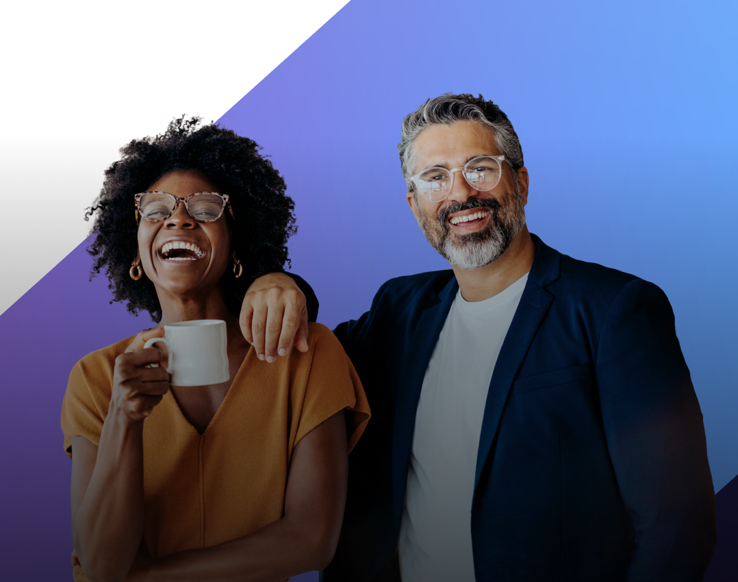 Two colleagues, a Black woman with glasses holding a mug and laughing, and a white man with glasses and a beard, stand together with his arm around her.
