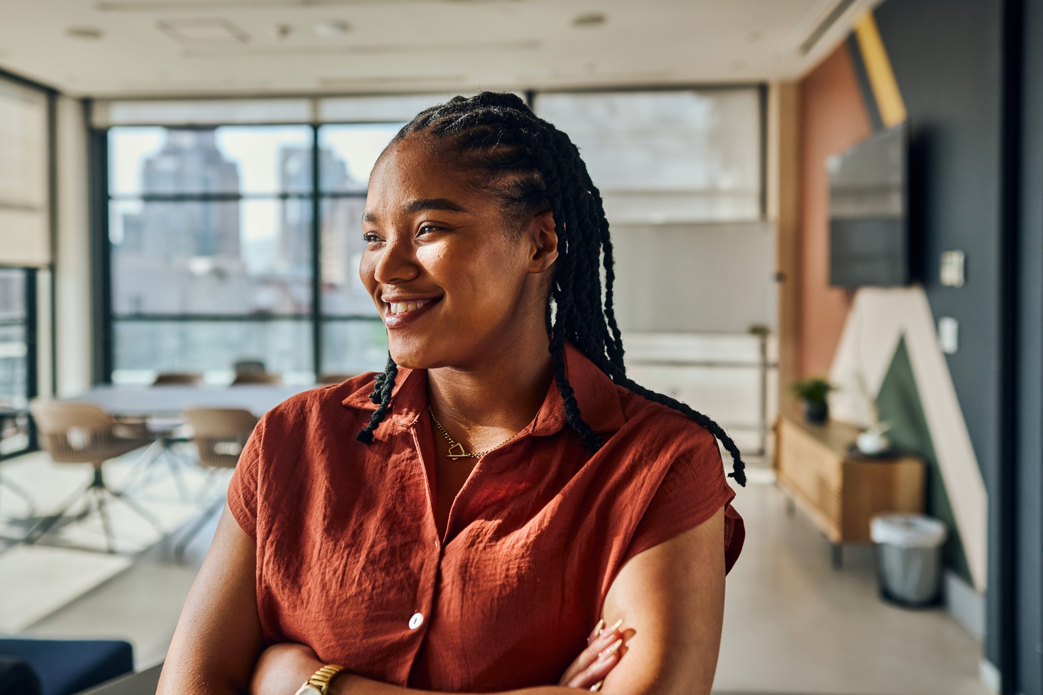 A smiling Black woman with braided hair wearing a rust-colored shirt in a bright, modern office.