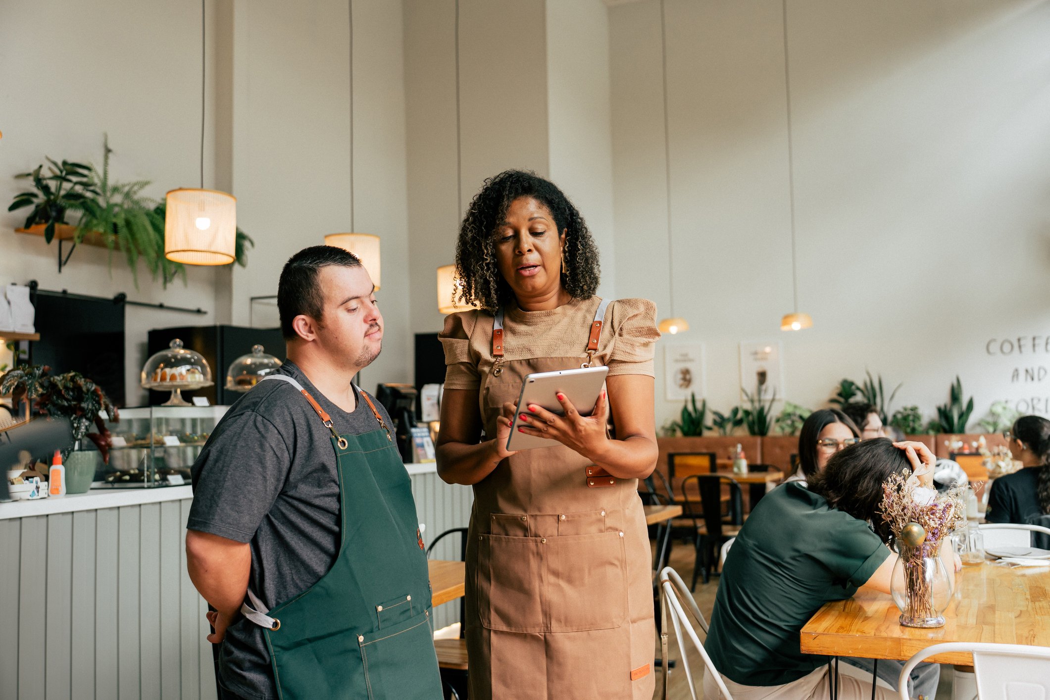 A woman in a brown apron shows a tablet to a young man in a green apron in a coffee shop.