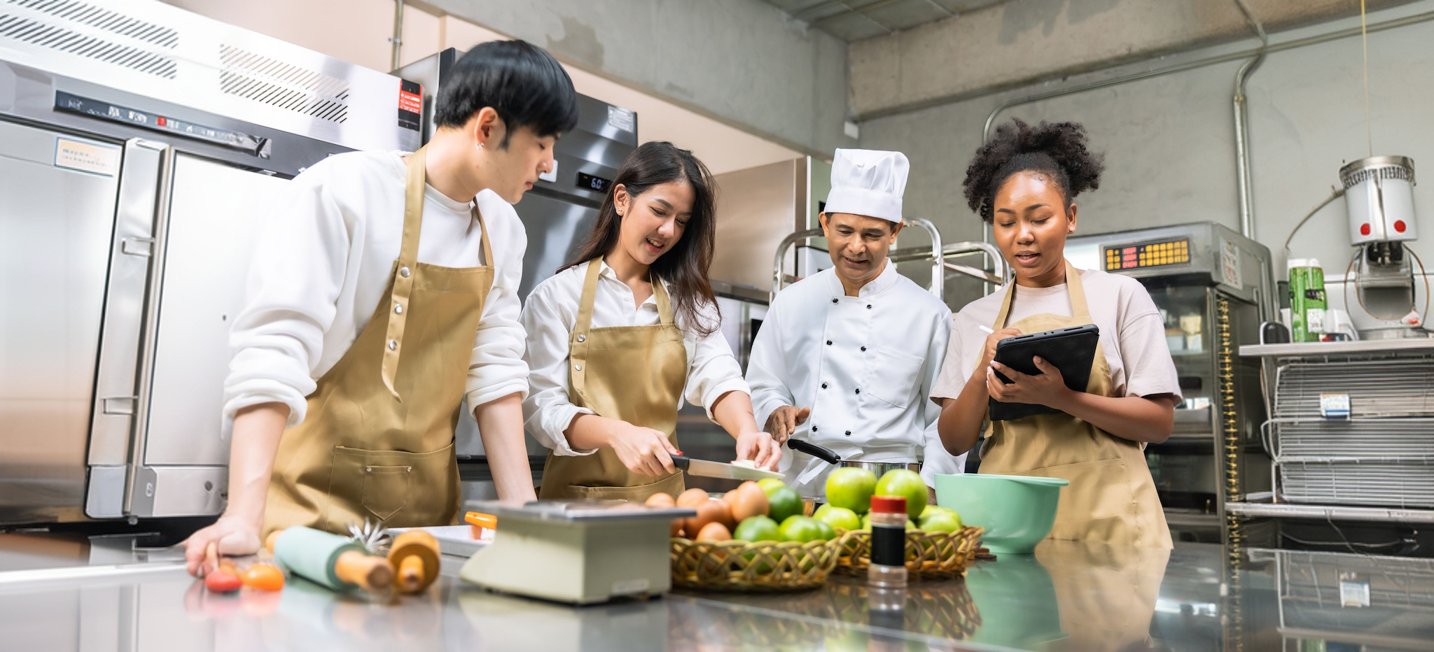 A professional chef instructing students in a kitchen while one student takes notes on a tablet.
