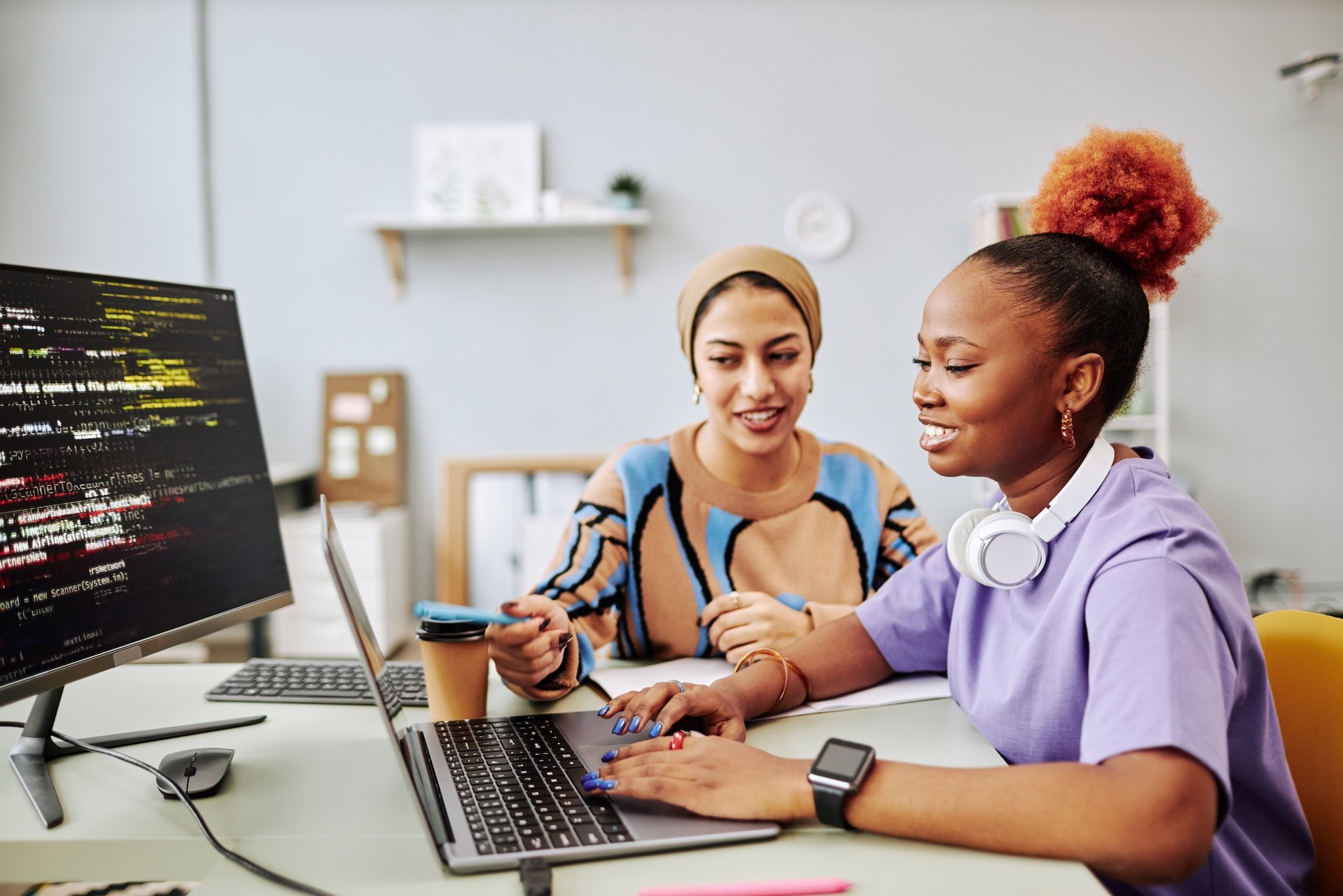 Two female students working together on a laptop and computer with code displayed on the screen.