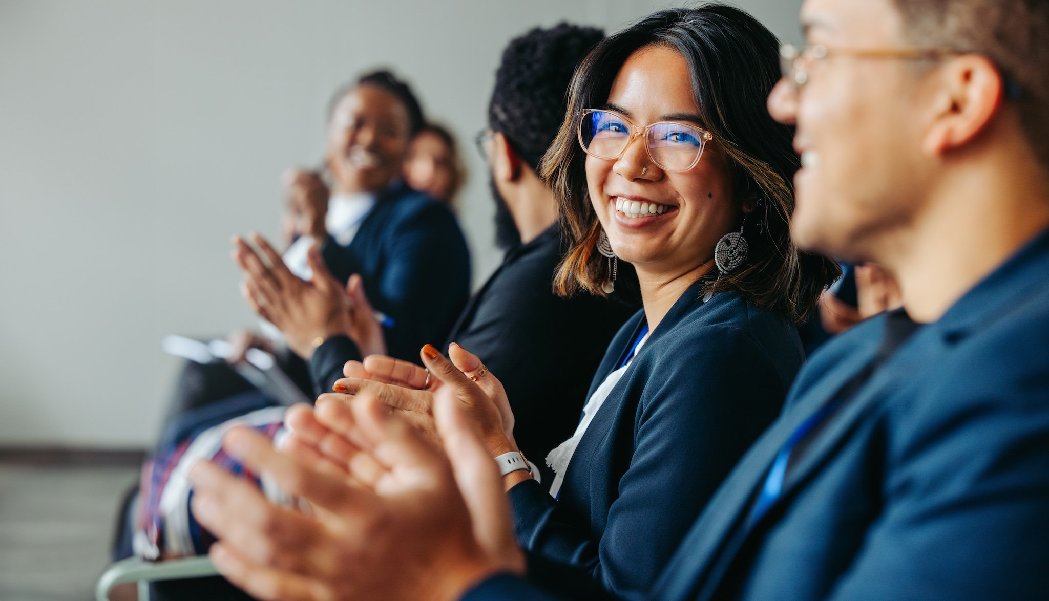 A diverse group of professionals in suits applauding at a conference, with a smiling woman in focus.