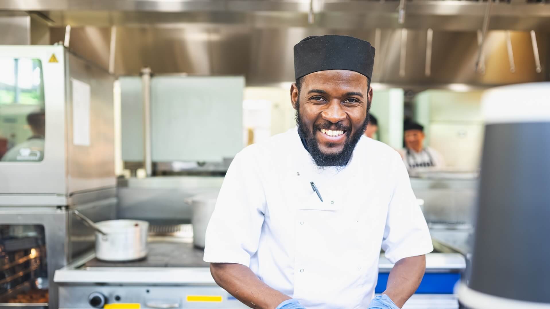 A smiling chef with a beard and black cap wearing a white uniform and blue gloves in a kitchen.
