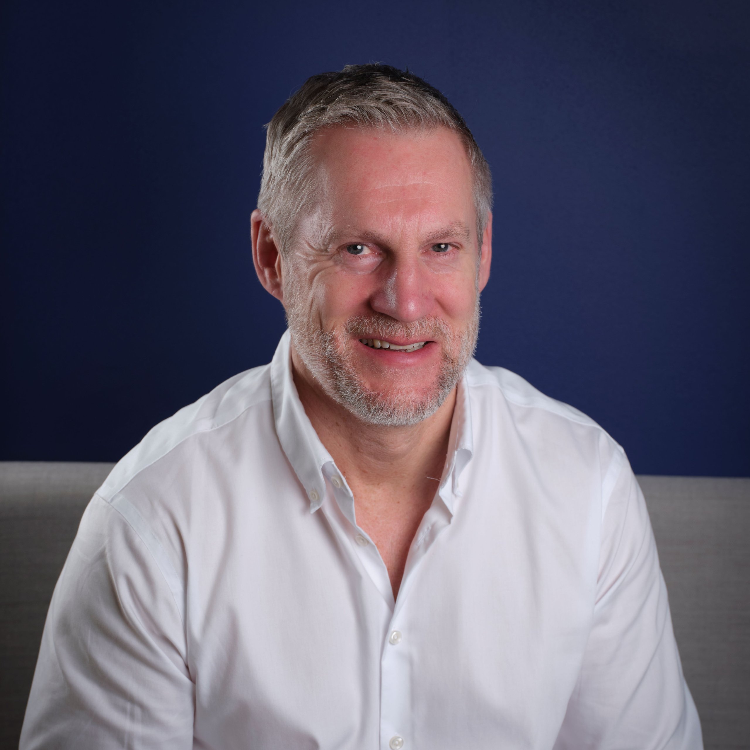 A middle-aged man with short grey hair and a beard, wearing a white shirt against a blue background.