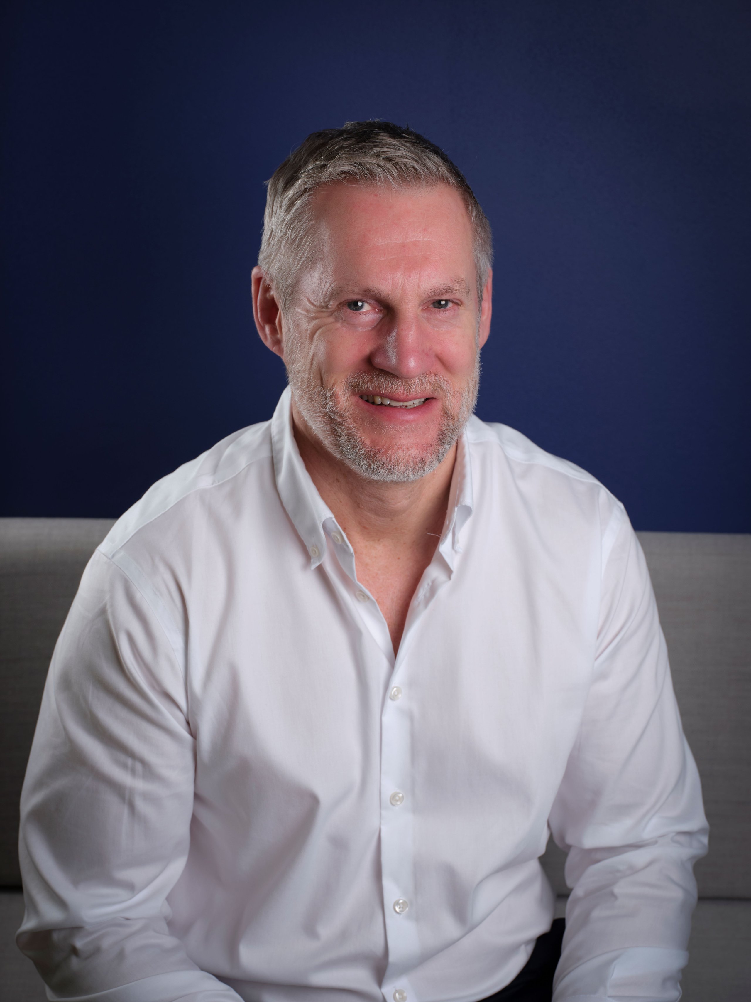A middle-aged man with short grey hair and a beard, wearing a white shirt against a blue background.