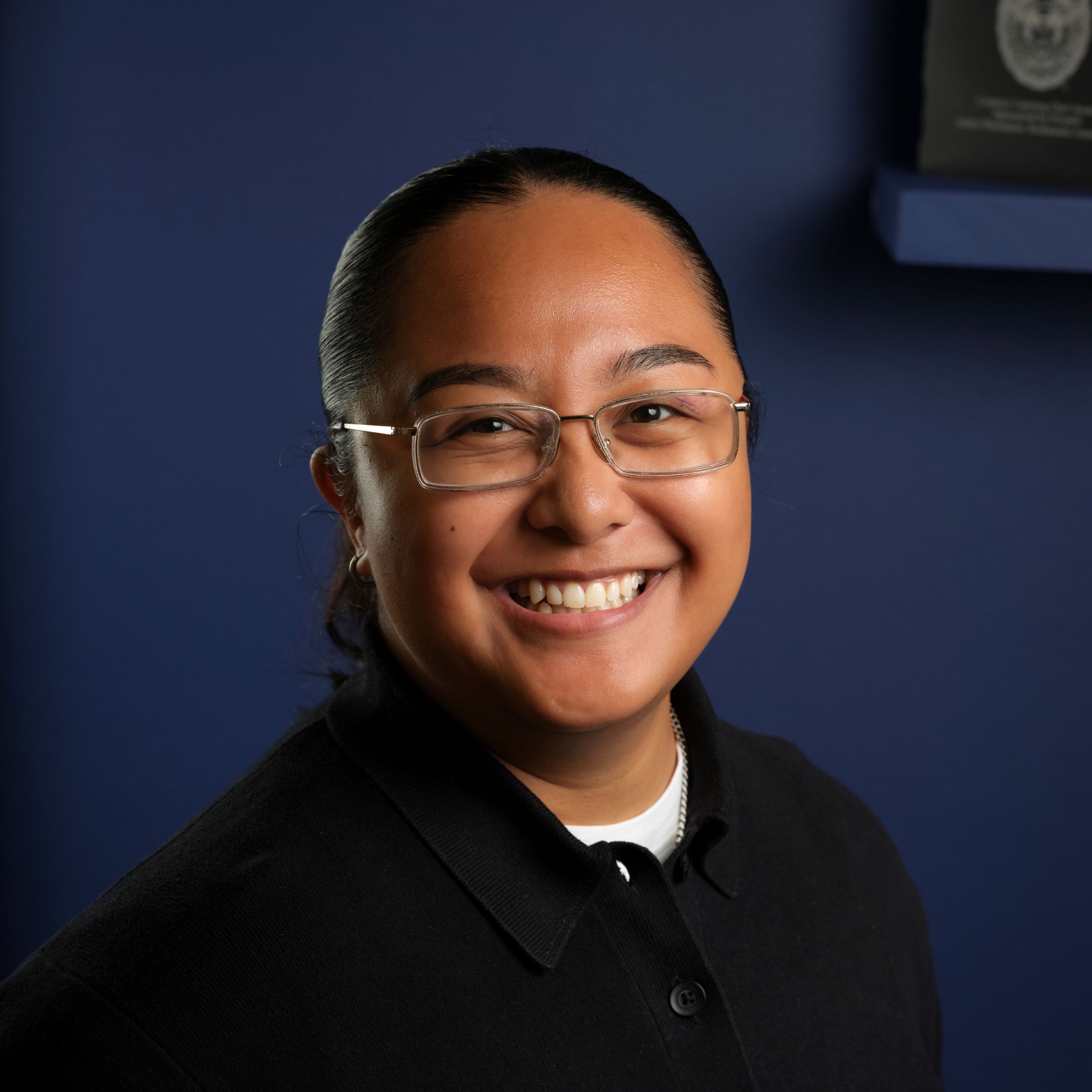 A smiling person with dark hair and glasses wearing a black polo shirt against a blue background.