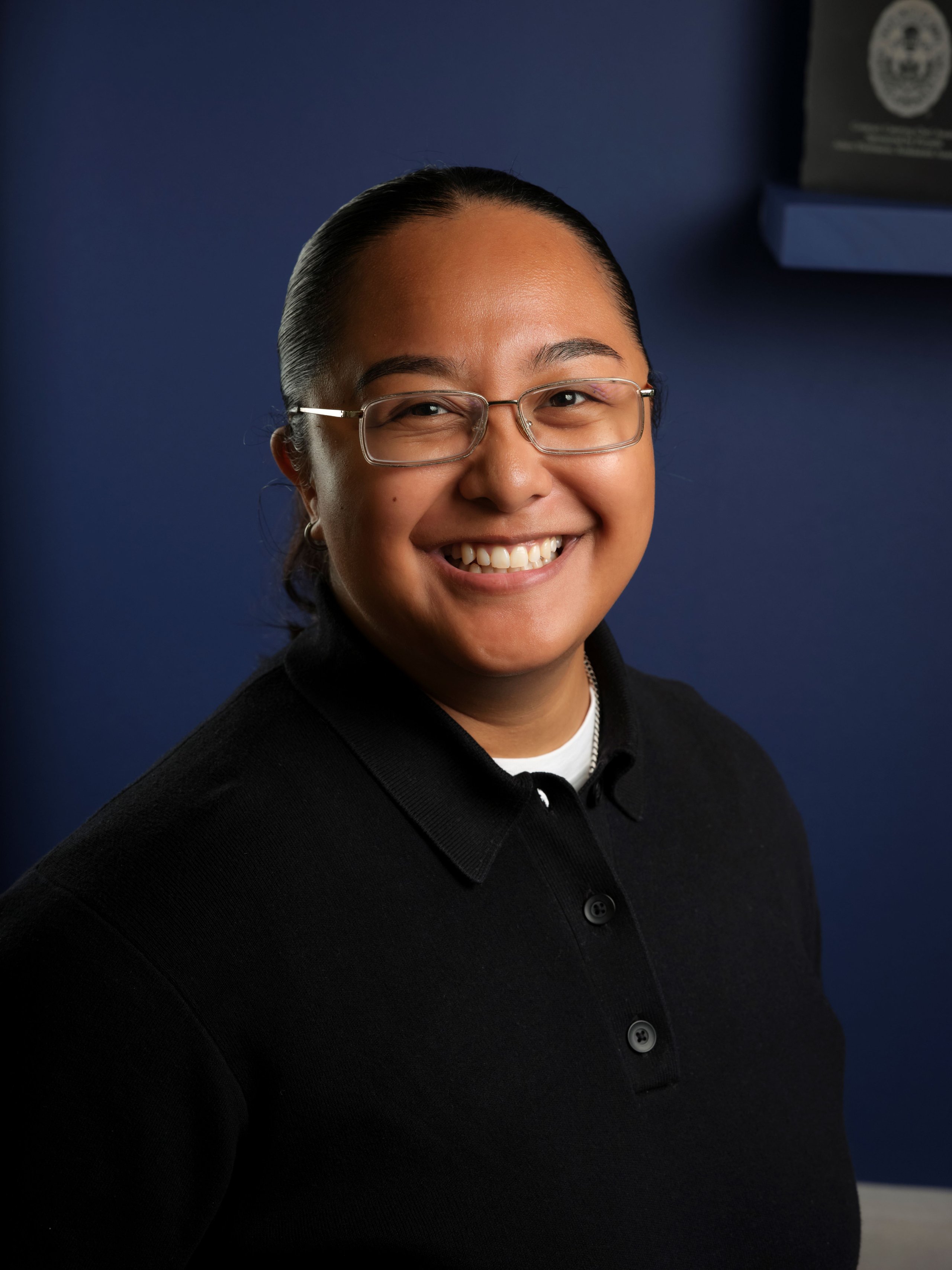 A smiling person with dark hair and glasses wearing a black polo shirt against a blue background.