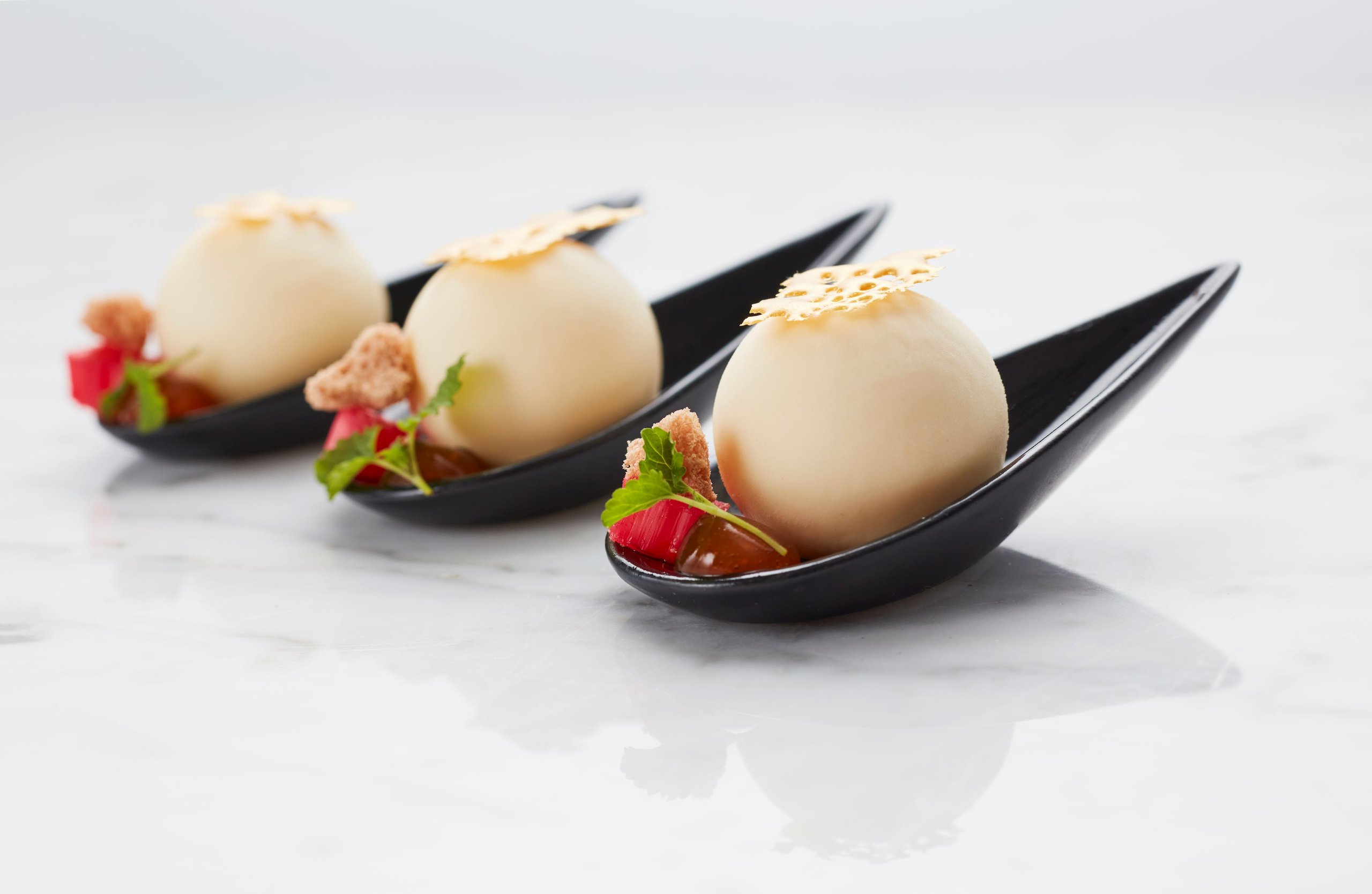 Three white chocolate spheres served on black tasting spoons with rhubarb, garnish, and a crisp.