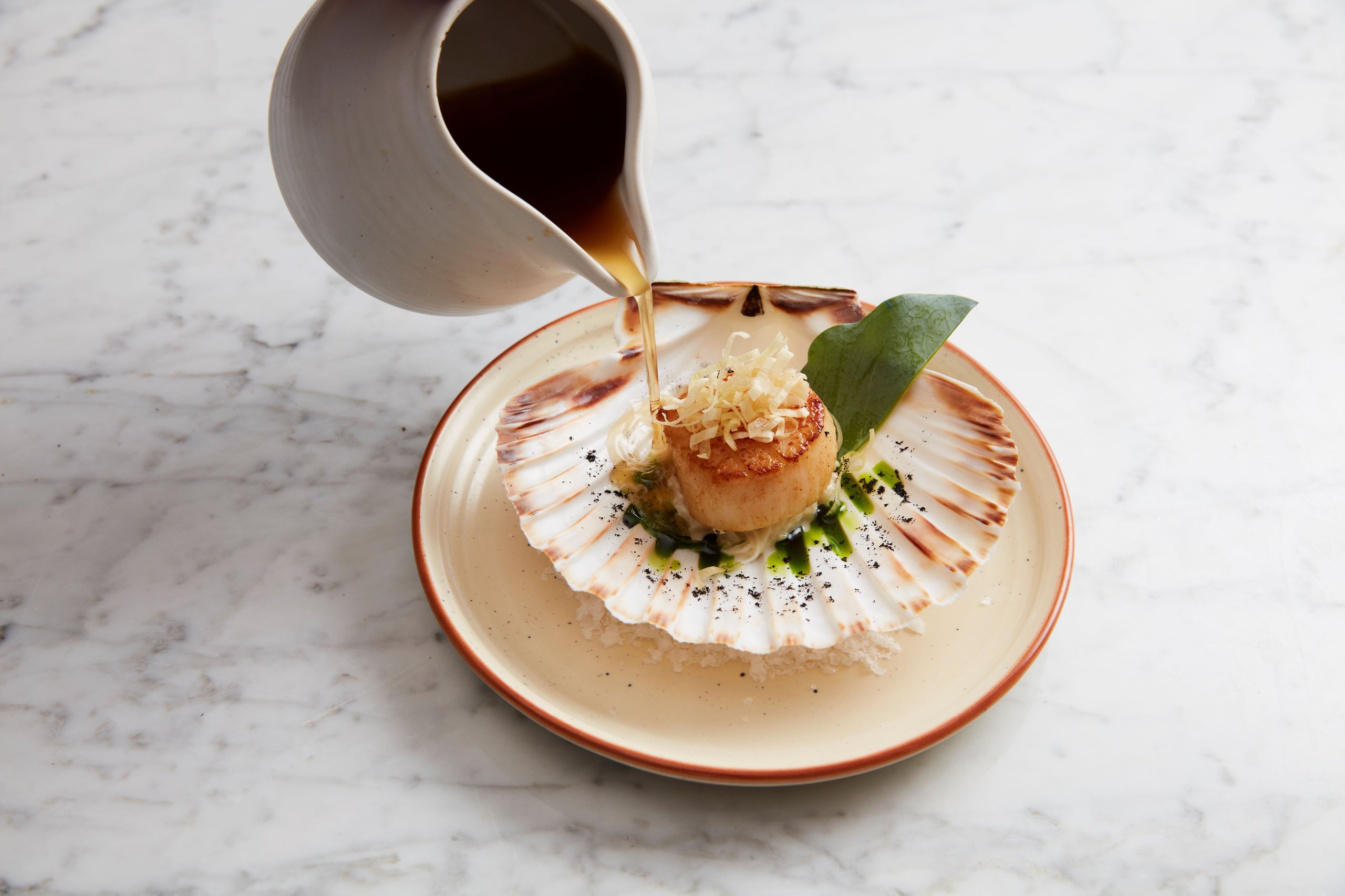 A seared scallop on a shell being garnished with broth poured from a ceramic pitcher.