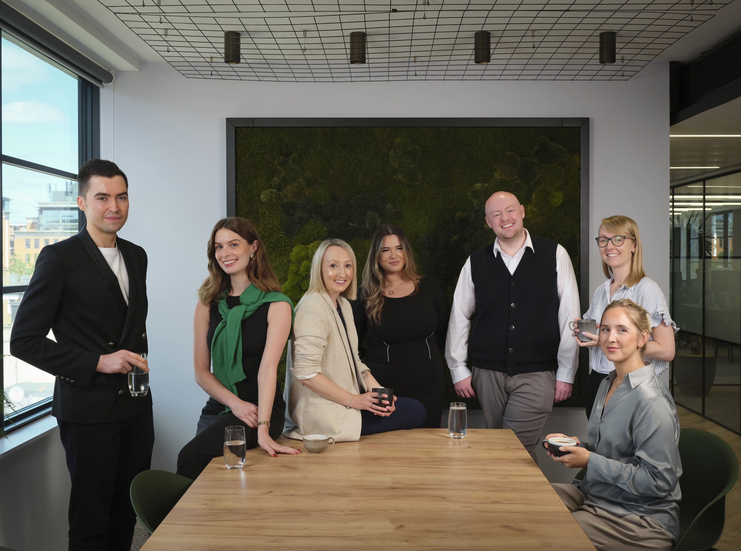 Group of seven colleagues smiling around a wooden table in a modern office with a moss wall.