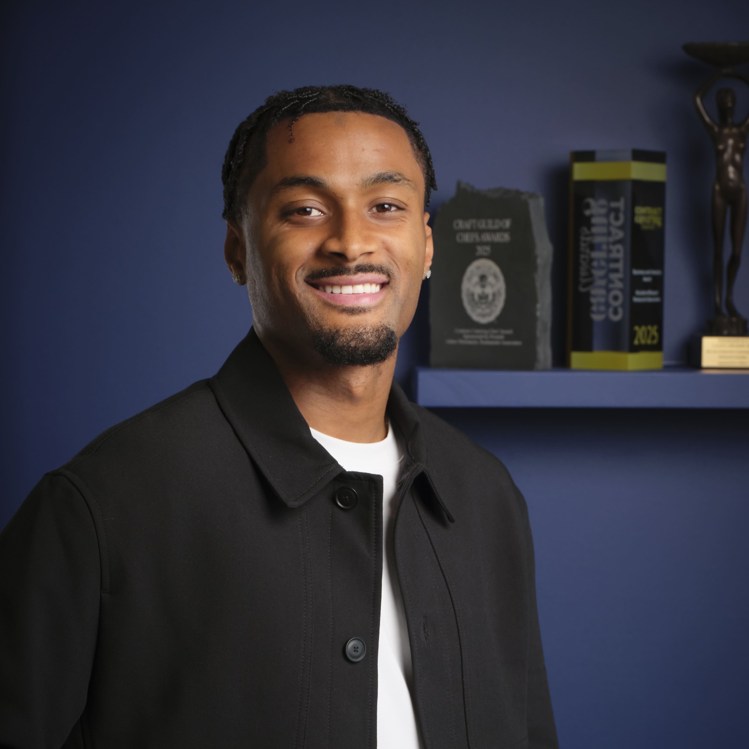 A smiling young man wearing a black collared jacket over a white shirt in front of an awards shelf.