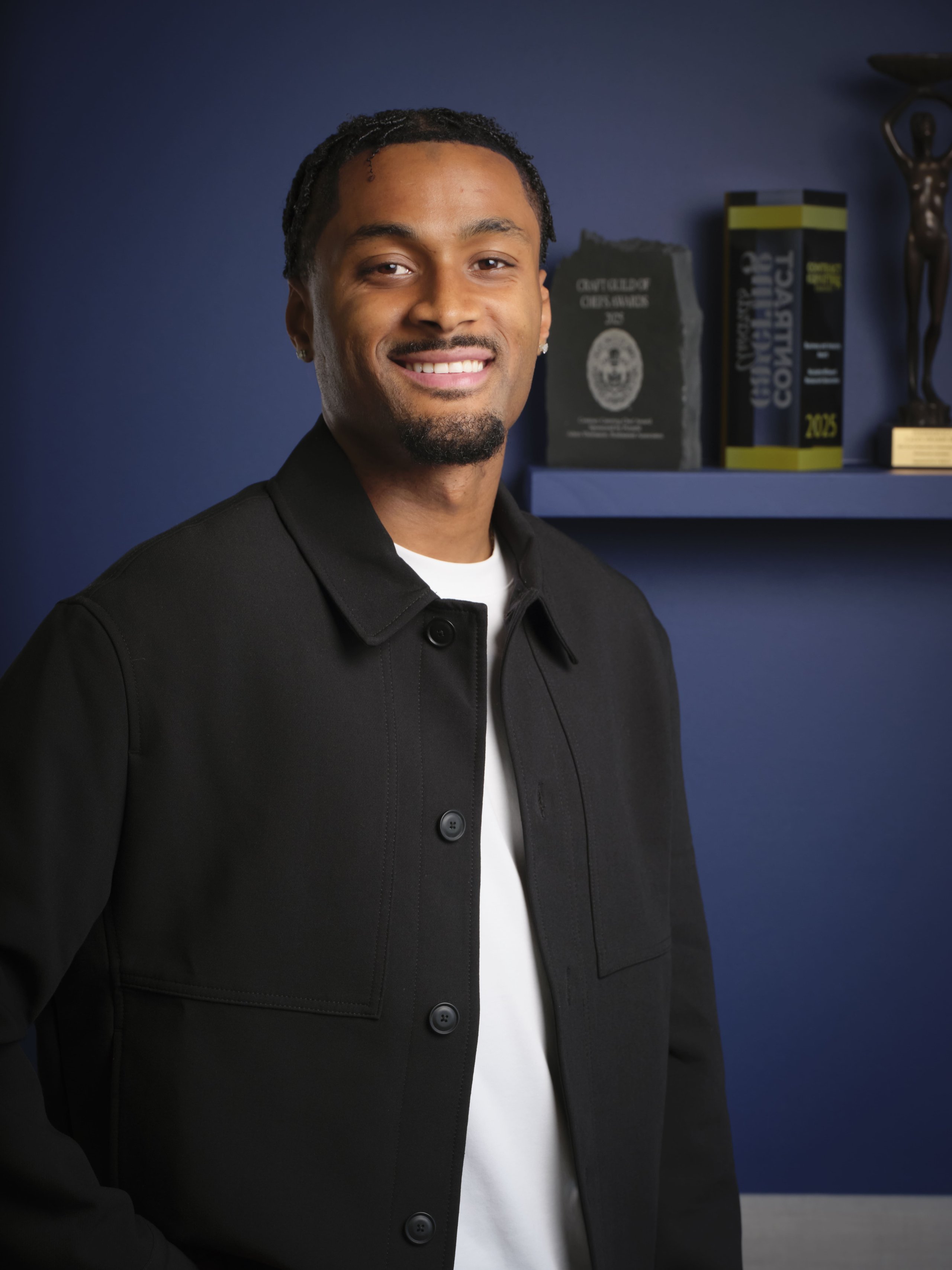 A smiling young man wearing a black collared jacket over a white shirt in front of an awards shelf.
