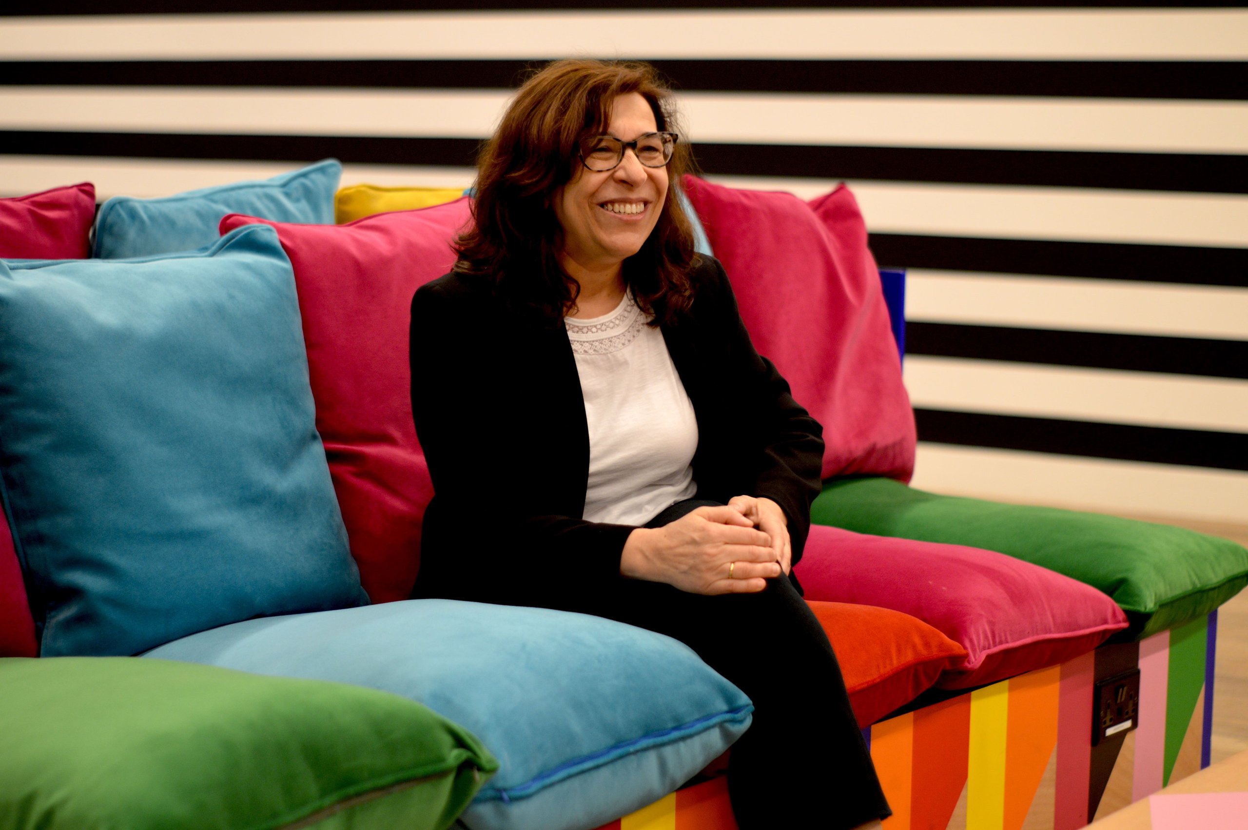 A woman in a black blazer and glasses sits smiling on a vibrant, multi-colored couch.