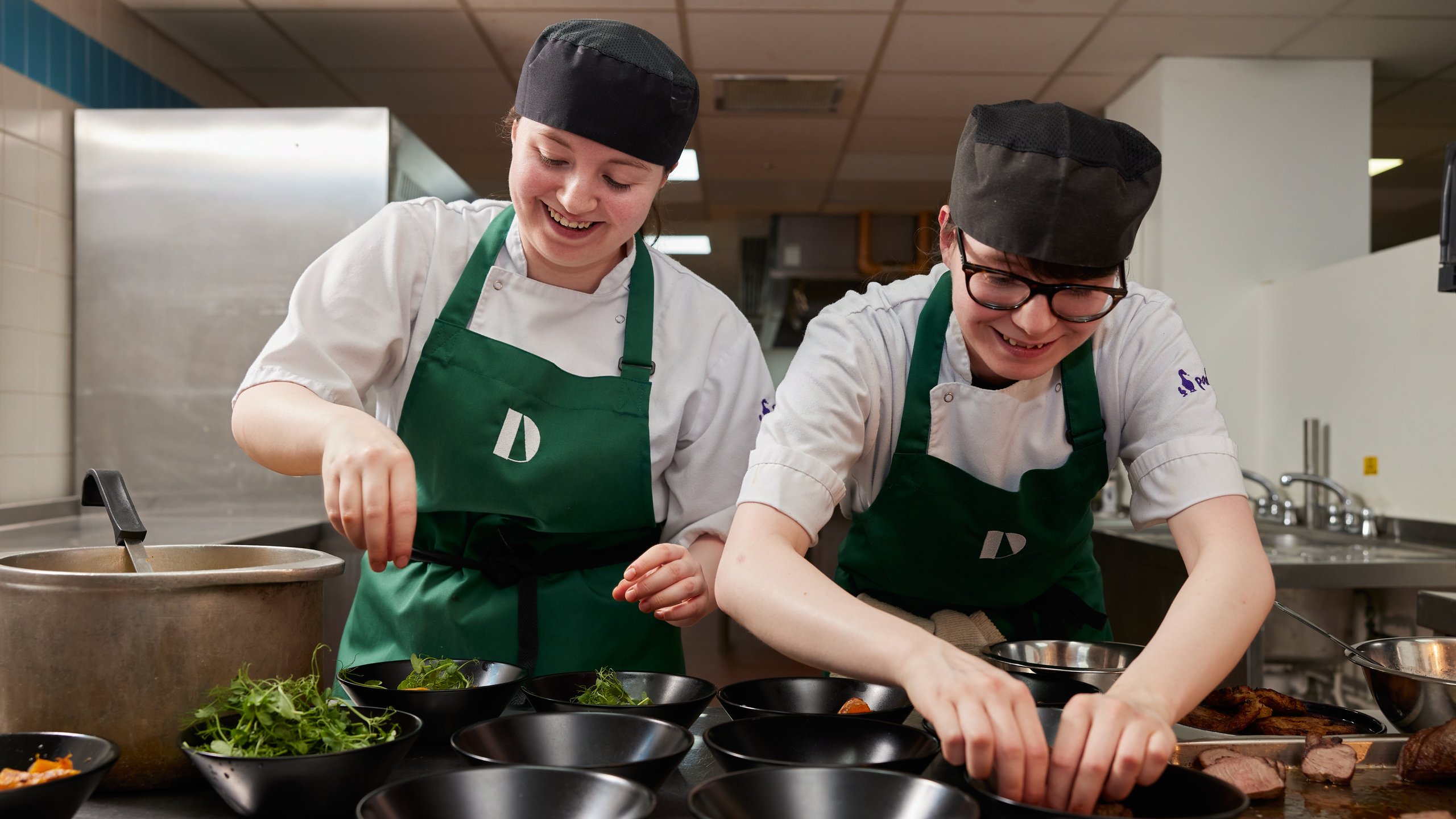 Two chefs in white uniforms and green aprons working together to plate food in a professional kitchen.